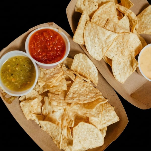 Two cardboard trays filled with tortilla chips, salsa, guacamole, and queso dip.