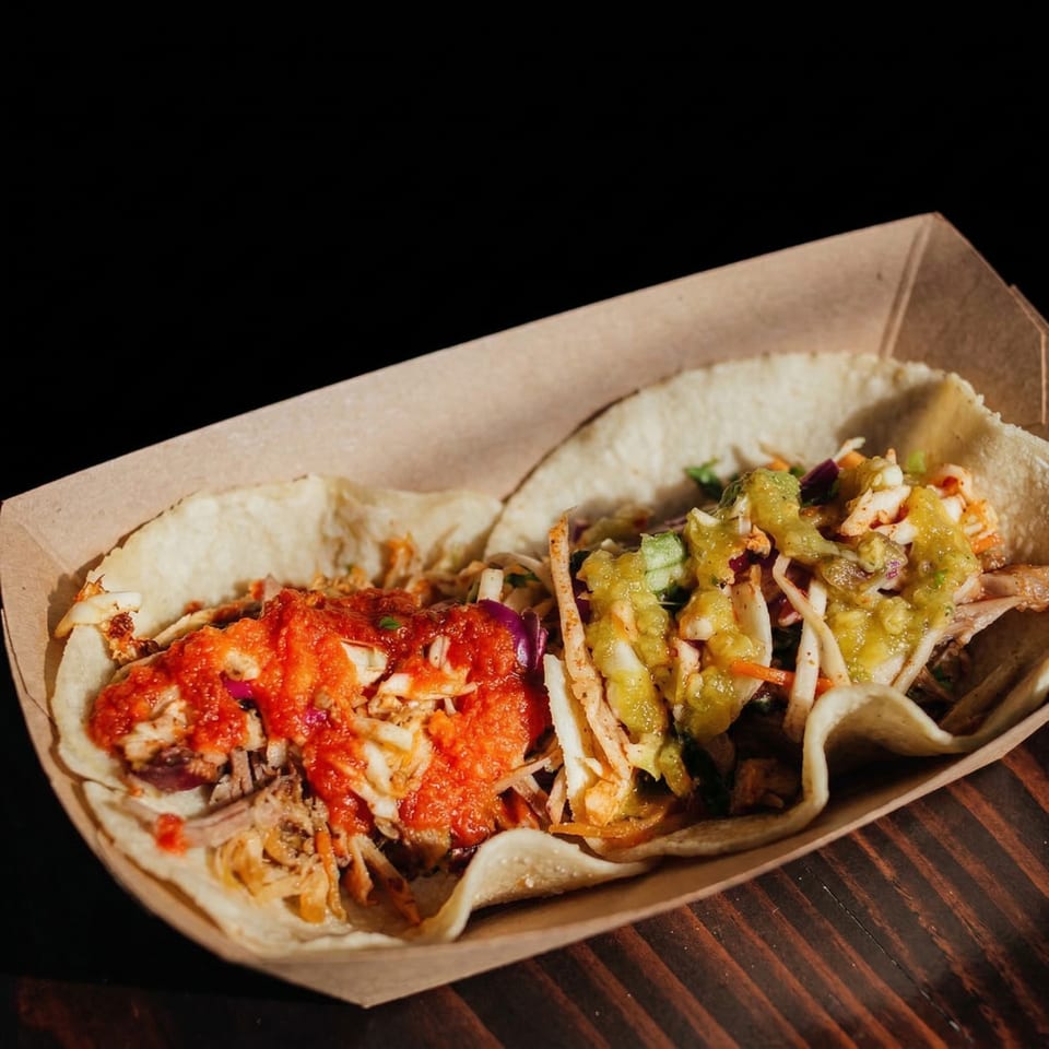 Two Muchachos tacos, one with red salsa and one with green salsa, in a cardboard tray.